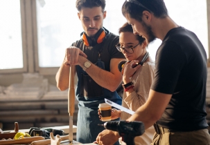 male carpenter showing something to coworker at his notebook papers in workshop